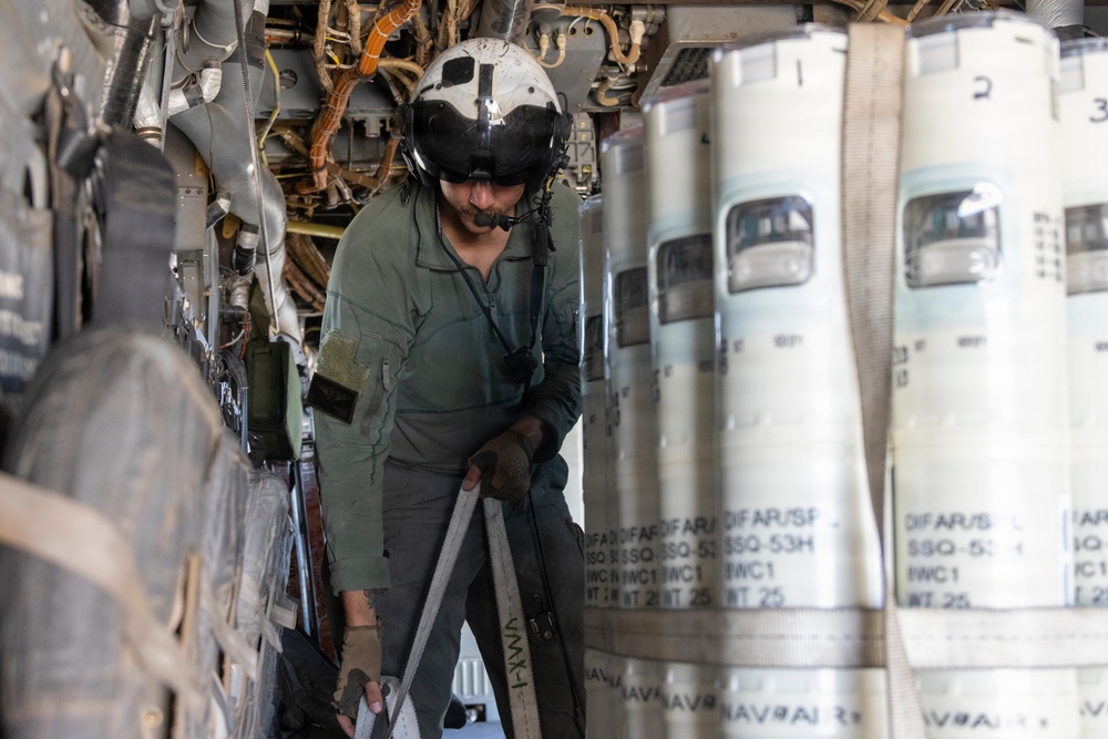 A military aircrew member wearing a flight helmet and olive drab flight suit is working inside what appears to be a maritime patrol or anti-submarine warfare aircraft. The individual is surrounded by rows of cylindrical sonobuoys secured with straps, identifiable by visible markings including 'SSQ-53H' and 'DIFAR' designations. The person appears to be handling or inspecting equipment within the aircraft's sonobuoy storage and launch bay.