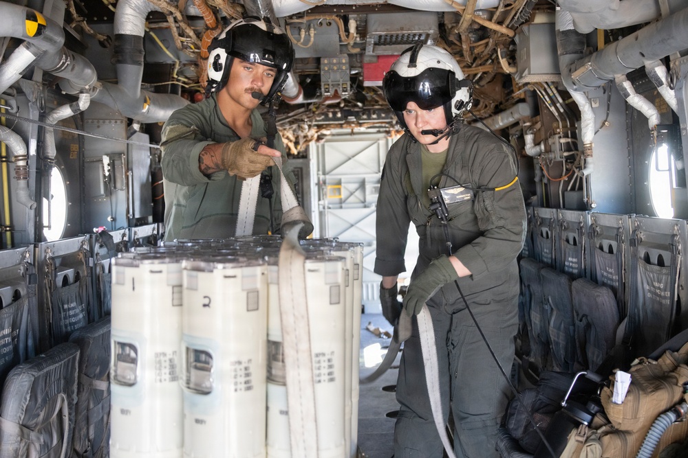 Two military aircrew members in flight suits and helmets are working inside what appears to be a large military helicopter, likely a CH-46 Sea Knight or CH-47 Chinook. They are handling what appears to be a bundled fire hose or similar equipment wrapped around cylindrical containers. The interior shows exposed mechanical systems, troop seating along the walls, and various cargo equipment.
