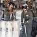 Two military aircrew members in flight suits and helmets are working inside what appears to be a large military helicopter, likely a CH-46 Sea Knight or CH-47 Chinook. They are handling what appears to be a bundled fire hose or similar equipment wrapped around cylindrical containers. The interior shows exposed mechanical systems, troop seating along the walls, and various cargo equipment.