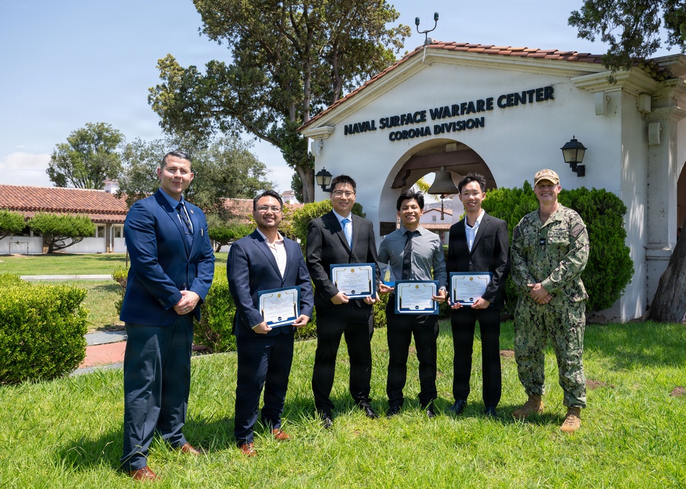 Six individuals pose for a group photo in front of the Naval Surface Warfare Center, Corona Division building. Four of the individuals are holding framed certificates or awards, suggesting a formal recognition ceremony. The group includes five civilians in business attire and one person in U.S. Navy military uniform.