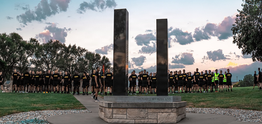 A large group of U.S. Army personnel dressed in black PT (physical training) uniforms are assembled around a memorial monument featuring two tall dark reflective pillars, likely representing the Twin Towers of the World Trade Center. The monument's base bears the inscription 'DONATED BY AUSA AND,' suggesting it was donated by the Association of the United States Army. The scene appears to be a 9/11 remembrance ceremony taking place at dusk or dawn on what appears to be a military installation.