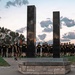 A large group of U.S. Army personnel dressed in black PT (physical training) uniforms are assembled around a memorial monument featuring two tall dark reflective pillars, likely representing the Twin Towers of the World Trade Center. The monument's base bears the inscription 'DONATED BY AUSA AND,' suggesting it was donated by the Association of the United States Army. The scene appears to be a 9/11 remembrance ceremony taking place at dusk or dawn on what appears to be a military installation.