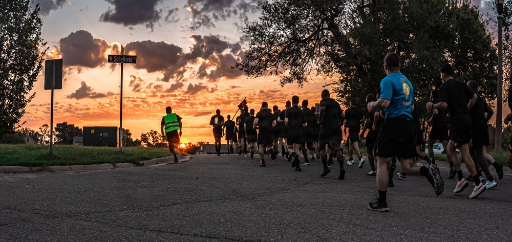 A large group of runners, likely military personnel based on their uniform black PT (physical training) attire, are running along a road at sunrise or sunset. A lead runner wearing a high-visibility green vest guides the formation, while another participant carries what appears to be a guidon or flag. The dramatic orange and purple sky creates a striking silhouette effect against the warm horizon.