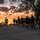 Victory Reception Company Welcome Run A large group of runners, likely military personnel based on their uniform black PT (physical training) attire, are running along a road at sunrise or sunset. A lead runner wearing a high-visibility green vest guides the formation, while another participant carries what appears to be a guidon or flag. The dramatic orange and purple sky creates a striking silhouette effect against the warm horizon.