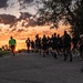 A large group of runners, likely military personnel based on their uniform black PT (physical training) attire, are running along a road at sunrise or sunset. A lead runner wearing a high-visibility green vest guides the formation, while another participant carries what appears to be a guidon or flag. The dramatic orange and purple sky creates a striking silhouette effect against the warm horizon.