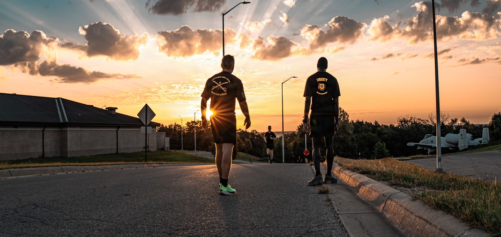 Two individuals wearing military-style black t-shirts walk along a road toward a dramatic sunset with sun rays bursting through clouds. One shirt reads 'LIBERTY' with an emblem, while the other displays crossed rifles insignia, suggesting a military unit affiliation. A static military aircraft is visible on the right side of the frame, and additional runners can be seen in the background.