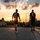 Victory Reception Company Welcome Run Two individuals wearing military-style black t-shirts walk along a road toward a dramatic sunset with sun rays bursting through clouds. One shirt reads 'LIBERTY' with an emblem, while the other displays crossed rifles insignia, suggesting a military unit affiliation. A static military aircraft is visible on the right side of the frame, and additional runners can be seen in the background.