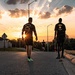 Two individuals wearing military-style black t-shirts walk along a road toward a dramatic sunset with sun rays bursting through clouds. One shirt reads 'LIBERTY' with an emblem, while the other displays crossed rifles insignia, suggesting a military unit affiliation. A static military aircraft is visible on the right side of the frame, and additional runners can be seen in the background.