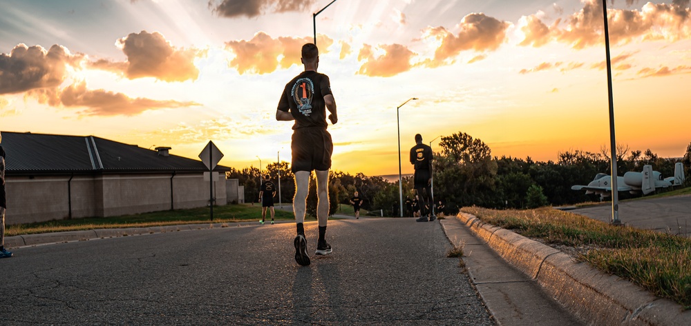 A group of runners in matching black athletic uniforms are running along a paved road during a dramatic sunset. The low-angle perspective emphasizes the lead runner in the foreground, with several others visible in the background. The setting appears to be a military installation, evidenced by aircraft displays and uniform attire visible in the background.