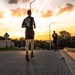 A group of runners in matching black athletic uniforms are running along a paved road during a dramatic sunset. The low-angle perspective emphasizes the lead runner in the foreground, with several others visible in the background. The setting appears to be a military installation, evidenced by aircraft displays and uniform attire visible in the background.