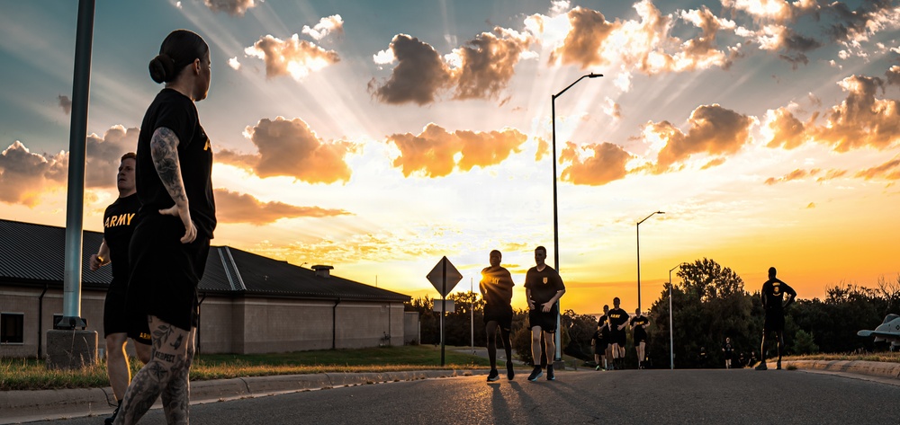 A group of U.S. Army soldiers in black PT (physical training) uniforms are running or exercising outdoors during a dramatic sunrise or sunset. The scene is set on a military installation road, with golden light rays breaking through clouds in the background. The foreground features a tattooed female soldier in an 'ARMY' shirt, with additional soldiers visible stretching into the distance.