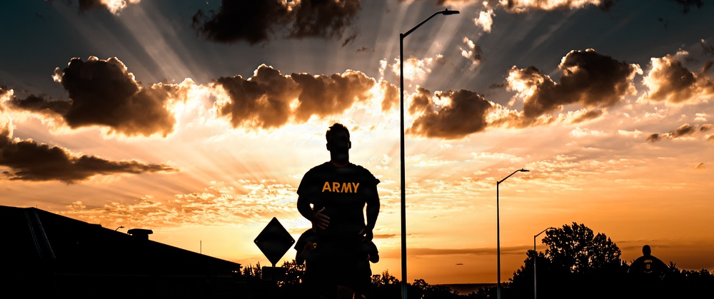 A silhouetted figure wearing an 'ARMY' t-shirt stands facing a dramatic sunset with crepuscular rays breaking through clouds. The scene is set along a road or street with lamp posts and signage visible in the background. The high-contrast backlighting creates a powerful, heroic composition.