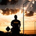 A silhouetted figure wearing an 'ARMY' t-shirt stands facing a dramatic sunset with crepuscular rays breaking through clouds. The scene is set along a road or street with lamp posts and signage visible in the background. The high-contrast backlighting creates a powerful, heroic composition.