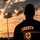 Victory Reception Company Welcome Run A silhouetted individual stands with their back to the camera, facing a dramatic sunset with rays of light breaking through clouds. The person is wearing a dark jacket with the word 'LIBERTY' and a crest/emblem on the back. The composition creates a powerful, contemplative mood with warm golden and orange tones dominating the sky.