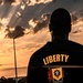 A silhouetted individual stands with their back to the camera, facing a dramatic sunset with rays of light breaking through clouds. The person is wearing a dark jacket with the word 'LIBERTY' and a crest/emblem on the back. The composition creates a powerful, contemplative mood with warm golden and orange tones dominating the sky.