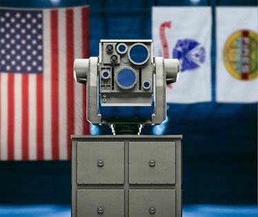 The image shows a military or defense-related optical/sensor device mounted on a gray metal cabinet with four drawers. The device features multiple circular lenses or apertures and appears to be on display in an official setting. An American flag and at least two other institutional flags are visible in the blurred background.