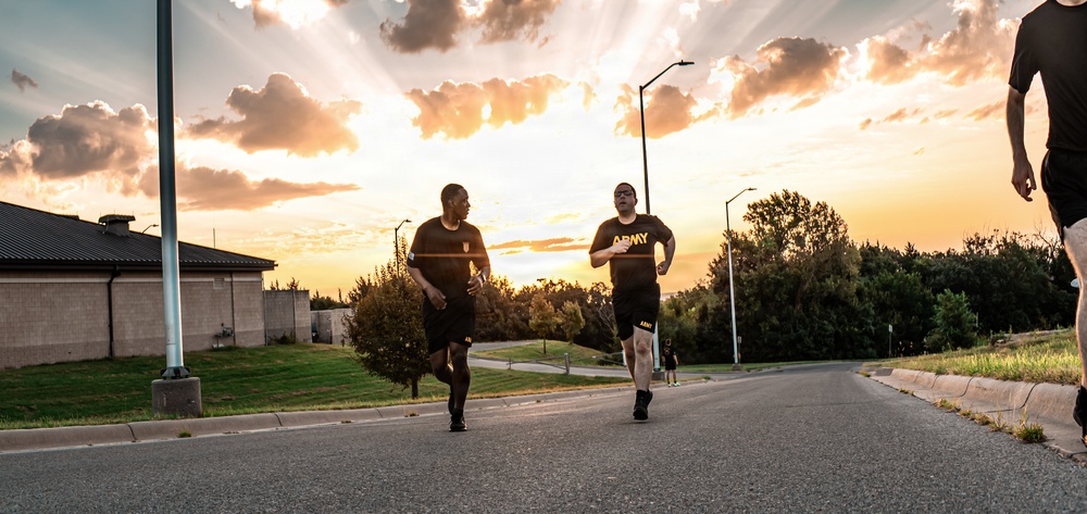 A group of U.S. Army soldiers are running on a paved road during what appears to be an early morning or evening physical training session. At least three individuals are visible wearing matching black Army PT (Physical Training) uniforms with gold 'ARMY' lettering. The scene is set against a dramatic sky with sunrays breaking through clouds, suggesting sunrise or sunset conditions.