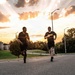 A group of U.S. Army soldiers are running on a paved road during what appears to be an early morning or evening physical training session. At least three individuals are visible wearing matching black Army PT (Physical Training) uniforms with gold 'ARMY' lettering. The scene is set against a dramatic sky with sunrays breaking through clouds, suggesting sunrise or sunset conditions.