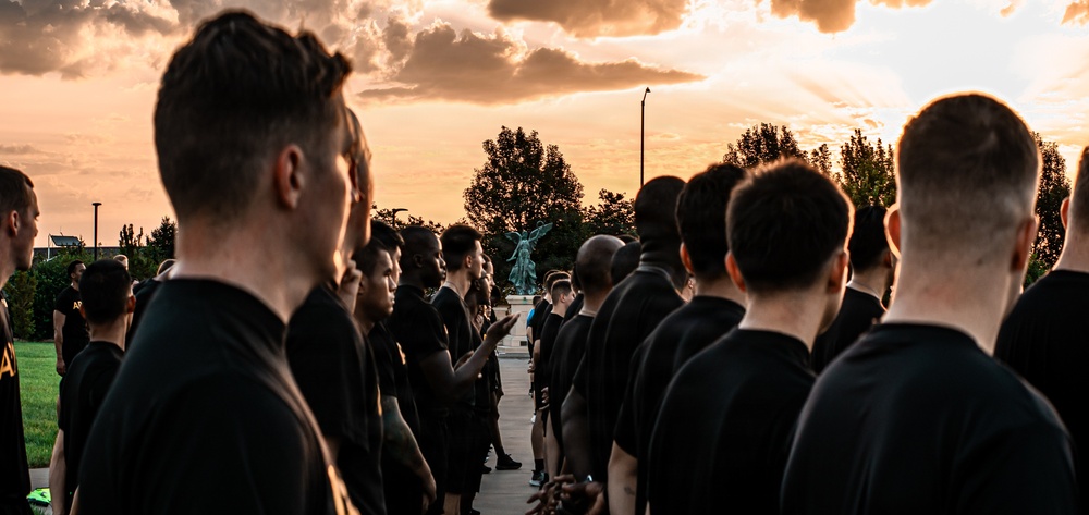 A large group of individuals dressed in matching black t-shirts are assembled outdoors at sunset or sunrise, standing in formation facing a bronze statue in the background. The dramatic orange and golden sky with clouds creates a striking backdrop. The uniformity of attire and organized formation suggests a military, ROTC, or structured group training event.