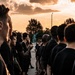 A large group of individuals dressed in matching black t-shirts are assembled outdoors at sunset or sunrise, standing in formation facing a bronze statue in the background. The dramatic orange and golden sky with clouds creates a striking backdrop. The uniformity of attire and organized formation suggests a military, ROTC, or structured group training event.