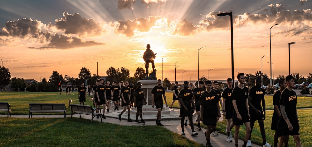 A group of U.S. Army soldiers or recruits in matching black 'ARMY' PT (physical training) uniforms walk along a paved path at sunset on what appears to be a military installation. Behind them stands a silhouetted soldier statue on a pedestal, dramatically backlit by golden sunrays breaking through clouds. The scene conveys military camaraderie, discipline, and patriotic symbolism.