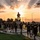 Victory Reception Company Welcome Run A group of U.S. Army soldiers or recruits in matching black 'ARMY' PT (physical training) uniforms walk along a paved path at sunset on what appears to be a military installation. Behind them stands a silhouetted soldier statue on a pedestal, dramatically backlit by golden sunrays breaking through clouds. The scene conveys military camaraderie, discipline, and patriotic symbolism.
