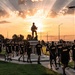 A group of U.S. Army soldiers or recruits in matching black 'ARMY' PT (physical training) uniforms walk along a paved path at sunset on what appears to be a military installation. Behind them stands a silhouetted soldier statue on a pedestal, dramatically backlit by golden sunrays breaking through clouds. The scene conveys military camaraderie, discipline, and patriotic symbolism.