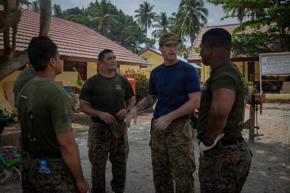 Four military personnel in uniform are gathered in an outdoor area near a building, engaged in a discussion or briefing. The setting appears to be a tropical location, likely in Southeast Asia, based on the palm trees, red-tiled roof structures, and a sign partially reading 'SD Negeri 12 MA' visible in the background. The individuals are wearing a mix of U.S. Marine Corps MARPAT camouflage and other military attire.
