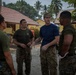 Four military personnel in uniform are gathered in an outdoor area near a building, engaged in a discussion or briefing. The setting appears to be a tropical location, likely in Southeast Asia, based on the palm trees, red-tiled roof structures, and a sign partially reading 'SD Negeri 12 MA' visible in the background. The individuals are wearing a mix of U.S. Marine Corps MARPAT camouflage and other military attire.