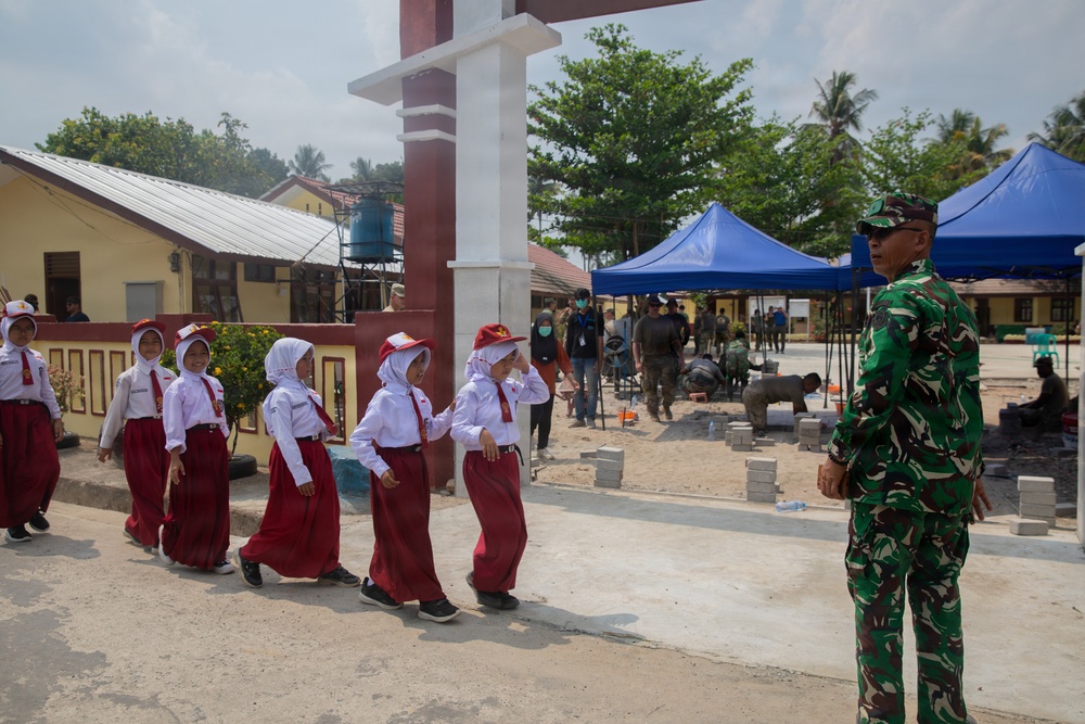 A group of young Indonesian schoolchildren in uniform are walking in a line past a military officer in camouflage uniform. The scene appears to take place at a school compound undergoing construction or renovation work, with workers visible in the background. One child appears to be saluting the military officer as they pass.