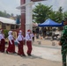 A group of young Indonesian schoolchildren in uniform are walking in a line past a military officer in camouflage uniform. The scene appears to take place at a school compound undergoing construction or renovation work, with workers visible in the background. One child appears to be saluting the military officer as they pass.