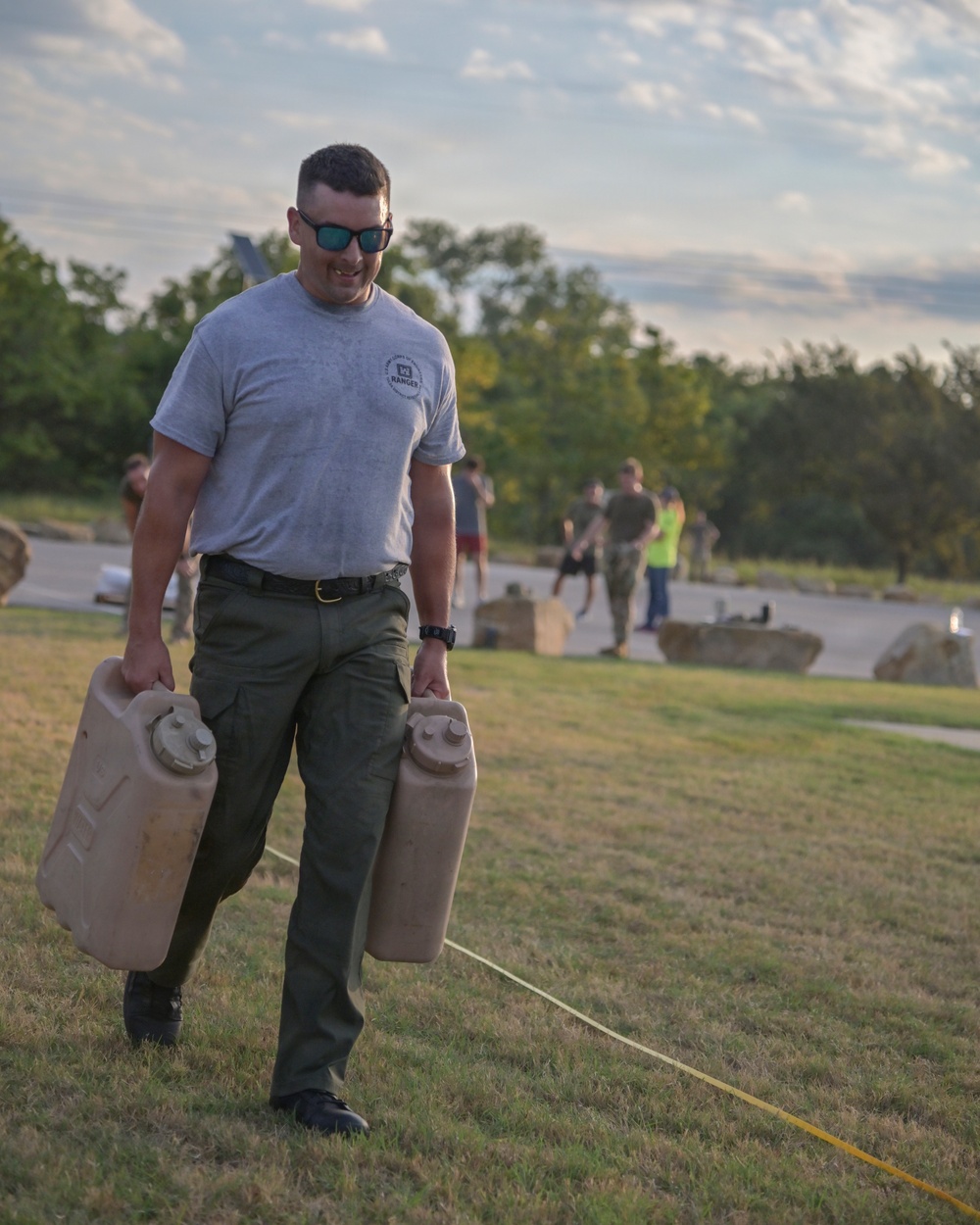 A man wearing a gray 'Ranger' t-shirt, olive green cargo pants, and sunglasses is carrying two large military-style jerry cans across a grassy field. He appears to be participating in a physical fitness or military-style challenge event, smiling as he walks. Several other participants and observers are visible in the background near a paved area.