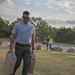 A man wearing a gray 'Ranger' t-shirt, olive green cargo pants, and sunglasses is carrying two large military-style jerry cans across a grassy field. He appears to be participating in a physical fitness or military-style challenge event, smiling as he walks. Several other participants and observers are visible in the background near a paved area.