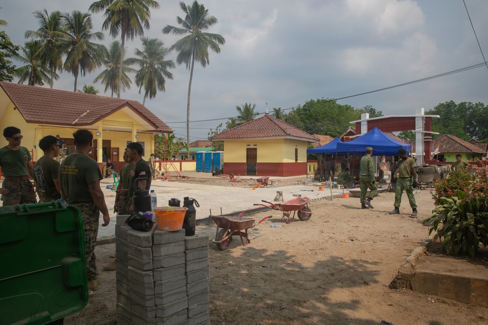 Military personnel in olive/green uniforms are engaged in construction or civic action work at what appears to be a village or compound in a tropical setting. Multiple soldiers are visible working around a partially completed paved area, with construction materials including concrete blocks, wheelbarrows, and tools present. The setting features yellow buildings with red-tiled roofs, palm trees, and a blue canopy tent in the background.