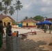 Military personnel in olive/green uniforms are engaged in construction or civic action work at what appears to be a village or compound in a tropical setting. Multiple soldiers are visible working around a partially completed paved area, with construction materials including concrete blocks, wheelbarrows, and tools present. The setting features yellow buildings with red-tiled roofs, palm trees, and a blue canopy tent in the background.