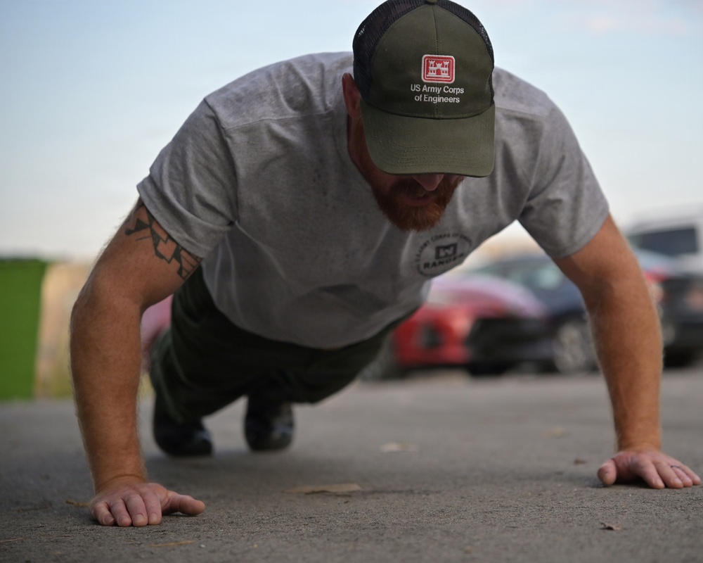 A man is performing push-ups on a paved outdoor surface. He is wearing a gray t-shirt and an olive green 'US Army Corps of Engineers' cap, with a visible tattoo on his left arm. The background shows a parking area with at least one red vehicle partially in frame.