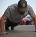 A man is performing push-ups on a paved outdoor surface. He is wearing a gray t-shirt and an olive green 'US Army Corps of Engineers' cap, with a visible tattoo on his left arm. The background shows a parking area with at least one red vehicle partially in frame.
