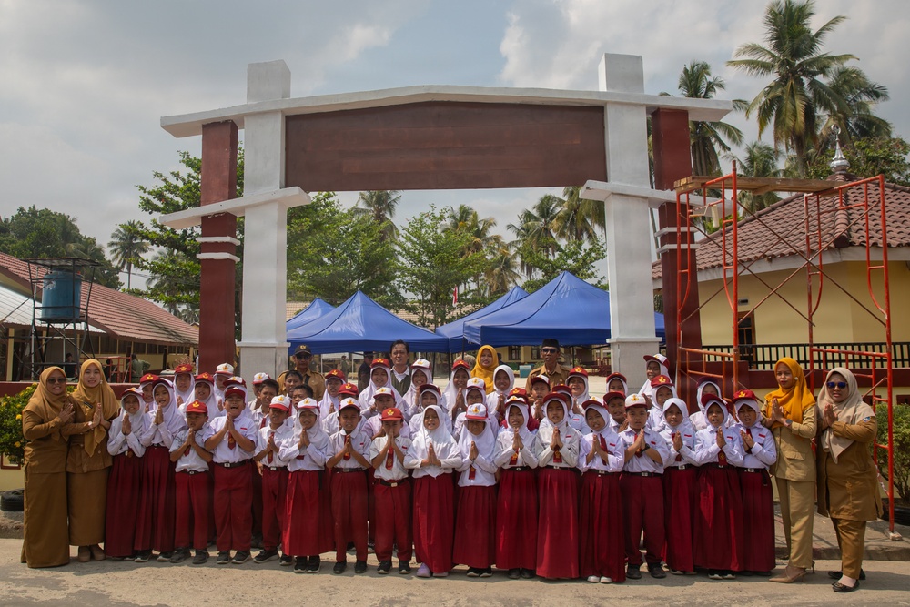A group of Indonesian primary school students in uniform pose for a group photo in front of a large decorative gate structure. The children are wearing white shirts with red skirts/pants and red caps, accompanied by several adult teachers or staff members. The setting appears to be a school compound with blue tents, palm trees, and buildings visible in the background.