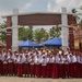 A group of Indonesian primary school students in uniform pose for a group photo in front of a large decorative gate structure. The children are wearing white shirts with red skirts/pants and red caps, accompanied by several adult teachers or staff members. The setting appears to be a school compound with blue tents, palm trees, and buildings visible in the background.