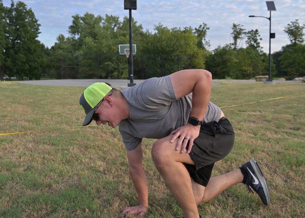 A man is kneeling on one knee on a grass field, bent forward with one hand on his thigh and the other touching the ground, appearing to be in pain or catching his breath. He is wearing a gray t-shirt, dark shorts, Nike sneakers, and a neon green trucker cap with sunglasses. A basketball court with a backboard is visible in the background.
