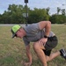 A man is kneeling on one knee on a grass field, bent forward with one hand on his thigh and the other touching the ground, appearing to be in pain or catching his breath. He is wearing a gray t-shirt, dark shorts, Nike sneakers, and a neon green trucker cap with sunglasses. A basketball court with a backboard is visible in the background.
