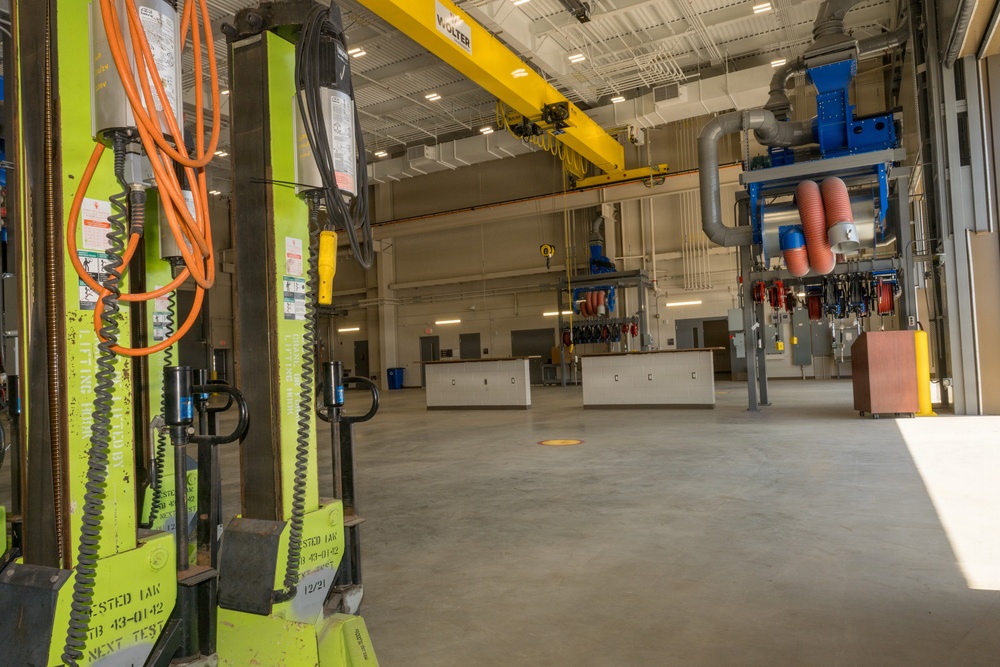 The image shows a large, modern industrial facility or maintenance workshop with high ceilings and a clean concrete floor. The space appears to be a well-equipped service bay or technical training facility featuring overhead crane systems, utility stations, and specialized equipment. The facility appears newly constructed or recently renovated, with organized workstations and industrial infrastructure.