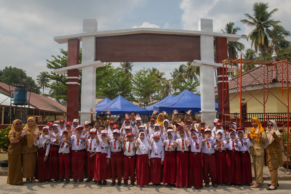 A large group of uniformed schoolchildren pose for a group photo in front of a decorative gate or entrance arch at what appears to be an Indonesian elementary school. The children are wearing matching white shirts with red bottoms and red caps, accompanied by several adult teachers or staff members. Many children are waving their hands enthusiastically toward the camera.