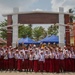 A large group of uniformed schoolchildren pose for a group photo in front of a decorative gate or entrance arch at what appears to be an Indonesian elementary school. The children are wearing matching white shirts with red bottoms and red caps, accompanied by several adult teachers or staff members. Many children are waving their hands enthusiastically toward the camera.