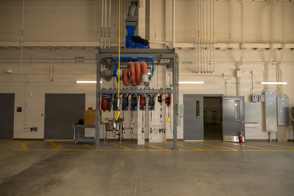 The image shows the interior of a large industrial or maintenance facility, likely a vehicle service bay or mechanical workshop. A central steel equipment tower/gantry holds multiple retractable hose reels, exhaust extraction hoses, and utility connections. The space appears newly constructed or recently outfitted, with clean concrete floors, organized utility runs along the ceiling, and minimal clutter.