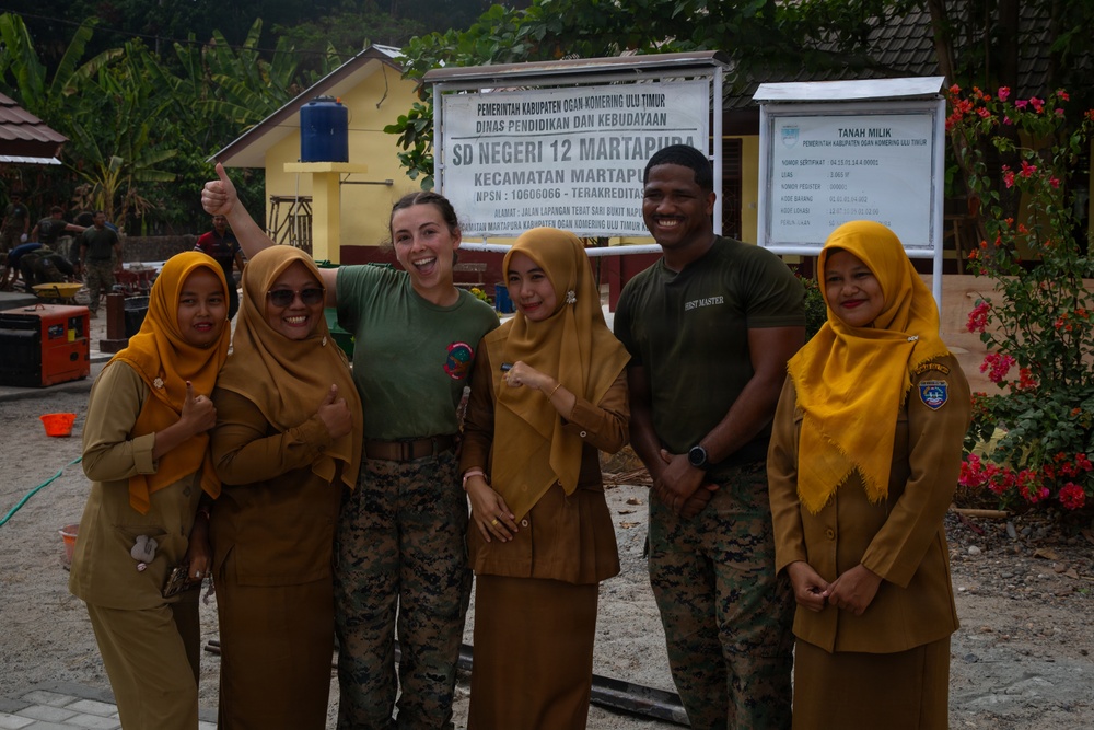 A group of six people pose for a photo in front of SD Negeri 12 Martapura school sign in Ogan Komering Ulu Timur, Indonesia. Two individuals in U.S. Marine Corps camouflage uniforms are posing with four local women wearing golden/mustard-colored hijabs and uniforms. The atmosphere is cheerful, with smiling faces and thumbs-up gestures.