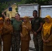 A group of six people pose for a photo in front of SD Negeri 12 Martapura school sign in Ogan Komering Ulu Timur, Indonesia. Two individuals in U.S. Marine Corps camouflage uniforms are posing with four local women wearing golden/mustard-colored hijabs and uniforms. The atmosphere is cheerful, with smiling faces and thumbs-up gestures.