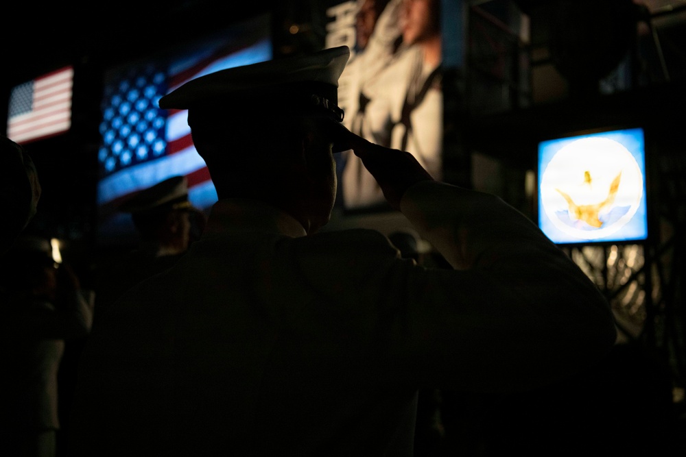 A silhouetted military service member in a peaked cap is captured mid-salute in a darkened environment. In the background, illuminated screens display an American flag and what appears to be a U.S. Navy emblem or seal. The dramatic low-light composition emphasizes the ceremonial nature of the moment.