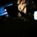 A silhouetted military service member in a peaked cap is captured mid-salute in a darkened environment. In the background, illuminated screens display an American flag and what appears to be a U.S. Navy emblem or seal. The dramatic low-light composition emphasizes the ceremonial nature of the moment.