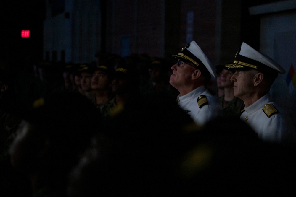 A group of military personnel are gathered in a dimly lit indoor or semi-outdoor setting, looking upward attentively. Two senior naval officers in white dress uniforms with gold insignia are prominently visible in the foreground, surrounded by other uniformed personnel in darker camouflage attire. The scene suggests a formal military ceremony or presentation taking place at night or in low-light conditions.
